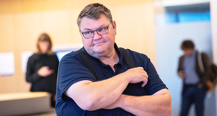 Han står med armarna i kors. Han har glasögon.