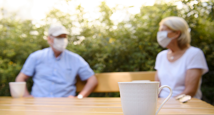 Två personer sitter en bit ifrån varandra på en bänk. DE har båda munskydd på sig. Framför dem syns ett bord. På bordet står en vit kaffekopp.