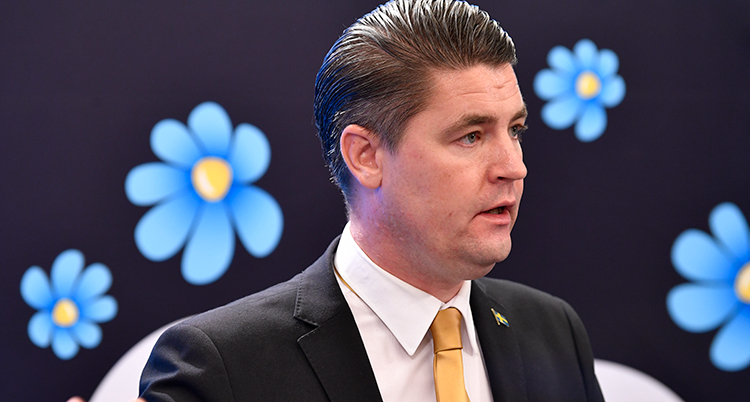 Närbild på Sjöstedt. Han ser allvarlig ut. I bakgrunden partiets symbol en blåklocka.