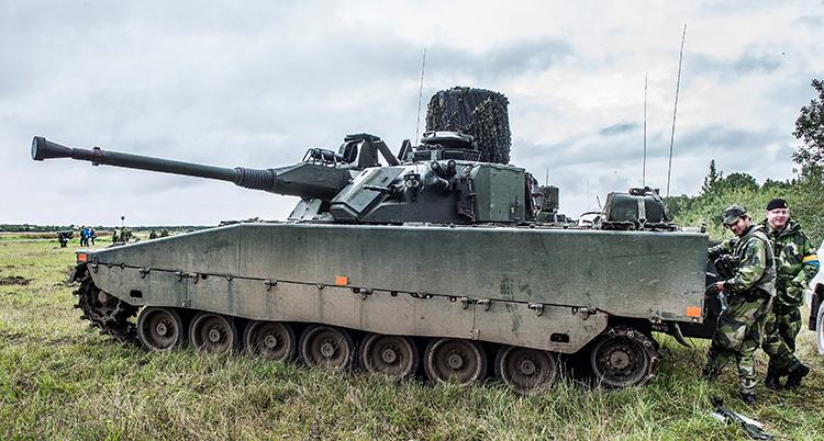 Några militärer står i gröna kläder runt ett stor stridsvagn. Den har flera hjul. De över på en stor gräsmatta.