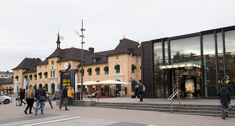 En bild på tågstationen i Uppsala. Till vänster syns ett gammalt ljust gult hus av sten och svart tak. Bredvid en byggnad av glas. Framför husen går några människor.