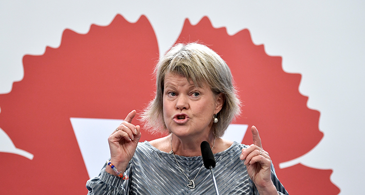 Hon talar. Hon har en mikrofon framför sig. I bakgrunden syns Vänsterpartiets märke i rött och vitt.