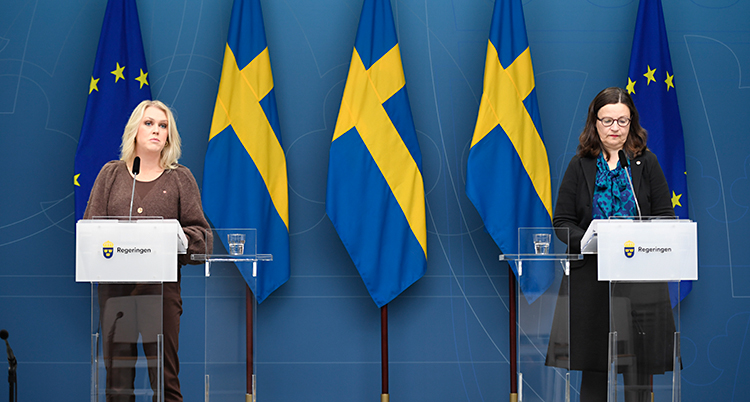 Två kvinnor står en scen. De pratar i varsin mikrofon. Lena Hallengren står till vänster och har ljust hår. Anna Ekström står till höger och har mörkt hår och glasögon. I bakgrunden finns Sveriges flagga och EUs flagga.