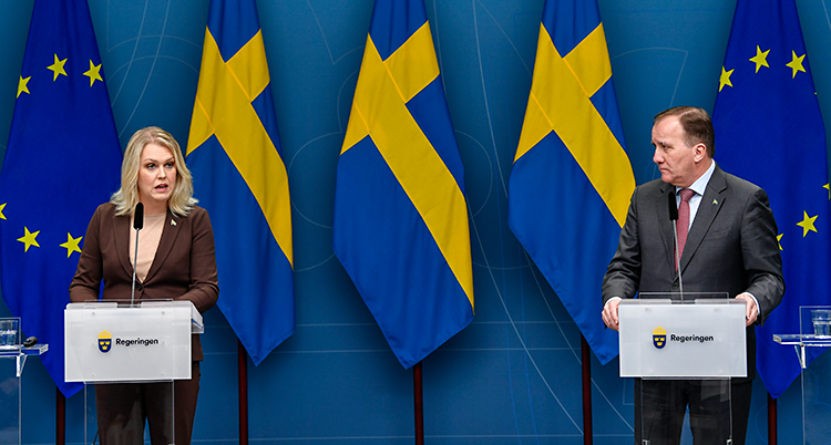 Ministern Lena Hallengren och statsminister Stefan Löfven pratar för journalister. De står på en scen och pratar i varsin mikrofon. I bakgrunden finns svenska flaggor och EUs flagga.