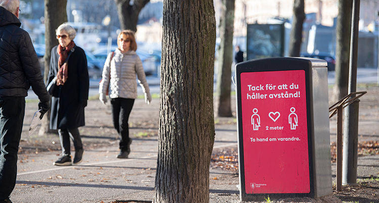 Två människor går utomhus bredvid dem syns en skylt som säger åt oss att hålla avstånd.