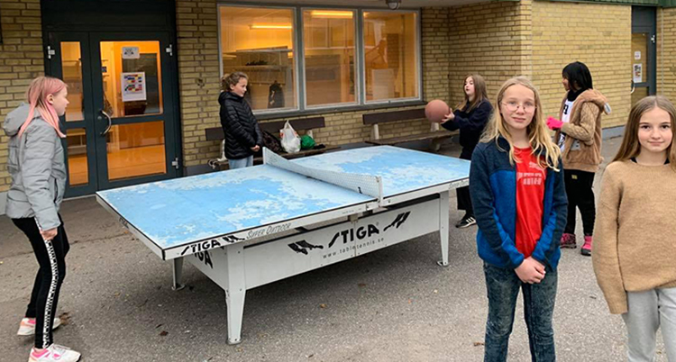 Flera barn spelar boll runt ett pingisbord utomhus. Två tjejer står vända mot kameran framför pingisbordet, den ena har ljus hår och blå jacka, den andra har brunt hår och beige tröja.