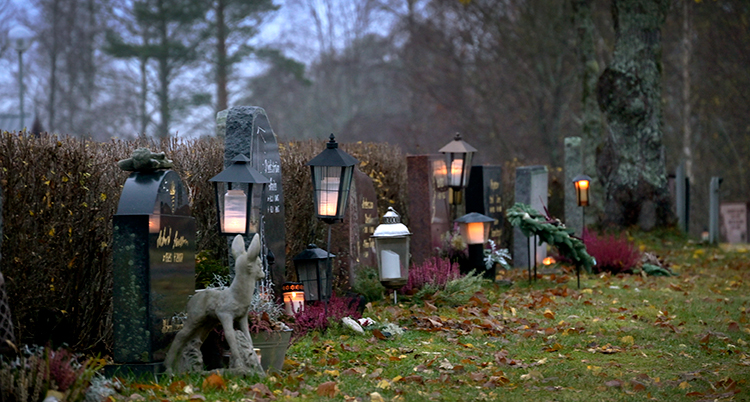 Flera stenar på en kyrkogård syns. De har alla en ljuslykta bredvid sig.