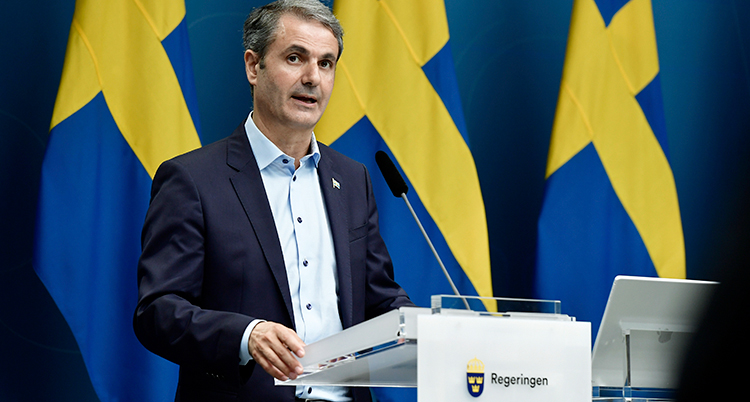 En man i grå kavaj och ljus skjorta står i en talarstol märkt med regeringen. Bakom syns tre svenska flaggor.