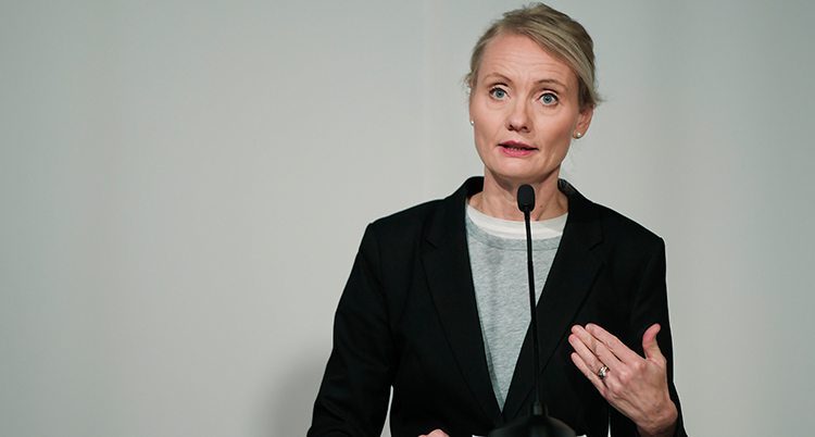 Hon talar i en mikrofon. Hon har svart kavaj på sig. Hon ser allvarlig ut.