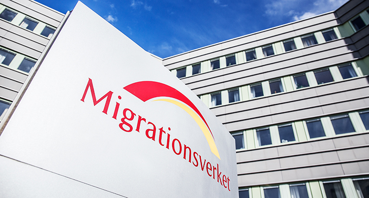 En skylt som det står Migrationsverket på i rött syns i förgrunden. Den är utomhus och syns mot en vit husfasad med många fönster och en blå himmel
