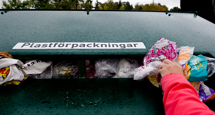 En hand slänger plast i en stor grön container för återvinning.