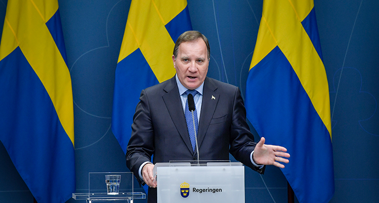 Han talar. Framför sig har han en mikrofon. Sveriges flagga syns i bakgrunden.
