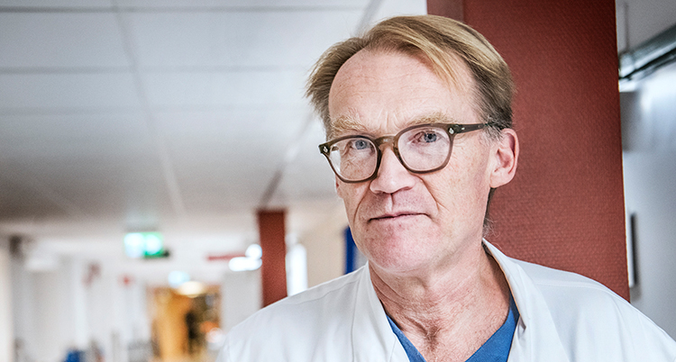 Han har vit rock på sig och glasögon. Han tittar in i kameran.