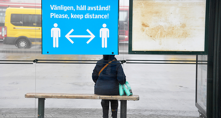 En person sitter ensam på en bänk i en busskur. På busskuren finns en stor skylt som visar att vi ska hålla avstånd.