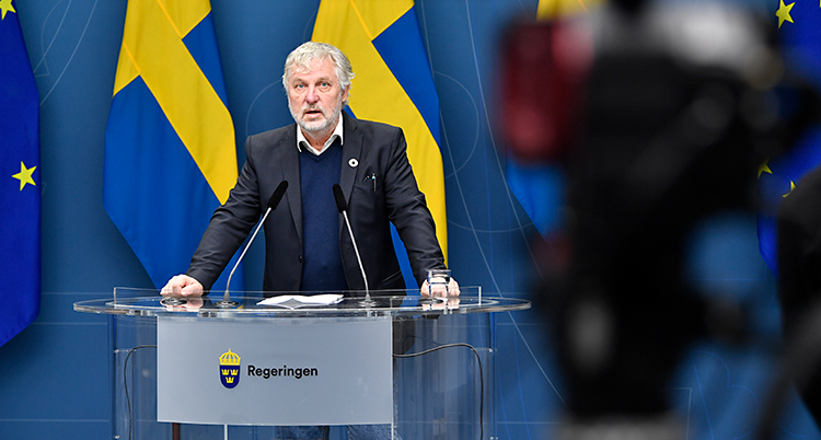 Peter Eriksson står och pratar i en mikrofon. Han står vid ett bord. I bakgrunden finns svenska flaggor. Han har grått hår och grått skägg. En kamera filmar honom.
