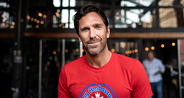 Henrik Lundqvist har en röd t-shirt. Han tittar in i kameran och ler litegrann. Han har brunt hår och skäggstubb.