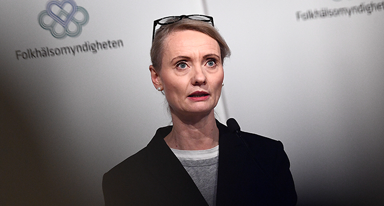 Karin Tegmark Wisell står och pratar i en mikrofon. Hon har blont hår och hon har sina glasögon uppe på huvudet. I bakgrunden finns en skärm där det står Folkhälsomyndigheten.