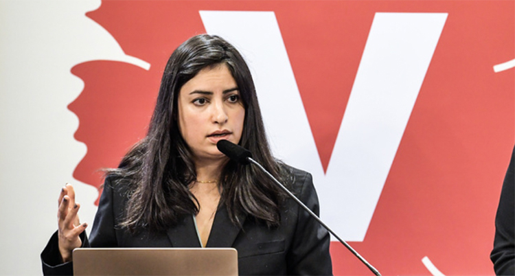 Hon står i en talarstol och pratar i en mikrofon. Bakom henne är partiets symbol ett V.