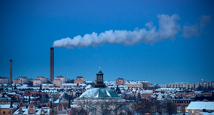 Det är en vy över Stockholm med många hus. En skorsten släpper ut rök mot en klarblå himmel.