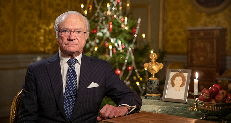 Kungen sitter i en stol och tittar in i kameran, han har blå kostym. Bakom finns en julgran.