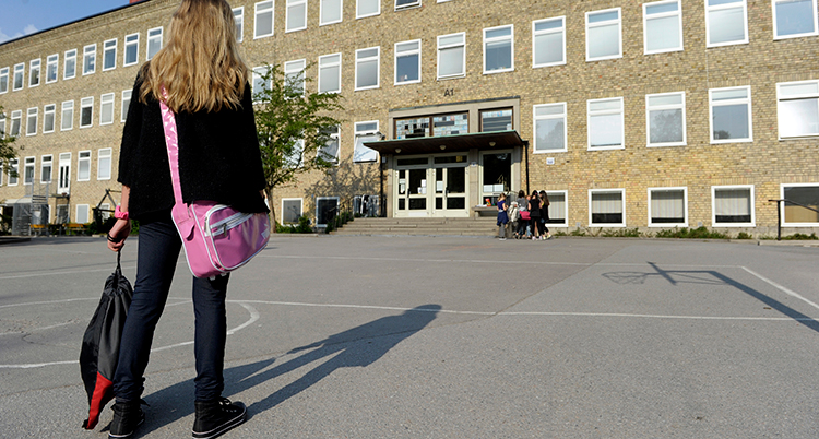 En flicka med en rosa skolväska över axeln och gympapåse i handen går över en asfalterad skolgård mot en skola i gult tegel