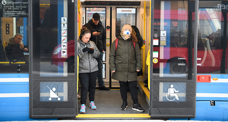 Två personer har klivit på en buss. Dörrarna har inte stängt sig ännu. En av personerna har munskydd.
