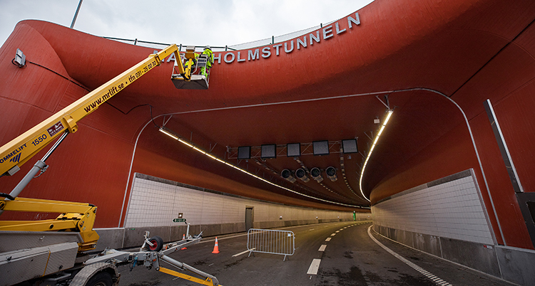 En röd biltunnel öppnar sig inåt, i förgrunden syns en gul kranarm. Svart asfalt leder in i tunneln.