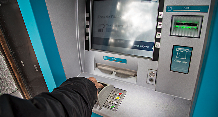 En person tar ut pengar från en bankomat. Vi ser bara armen på personen. Bankomaten har en skärm och olika knappar.