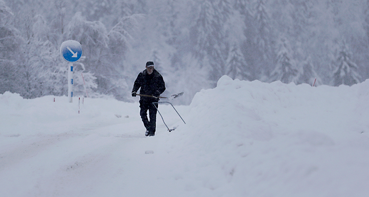 Bilden är tagen utomhus. Det är väldigt vitt. Det är massor av snö på en väg och bredvid vägen. En man går med en skovel och skottar. I bakgrunden syns en skog.