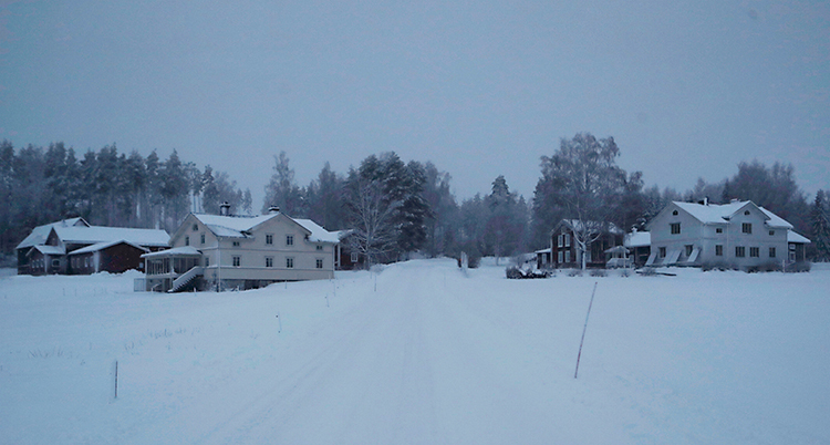 Bilden är tagen i en by på landet. Det är mycket snö på marken. Vi ser några hus. Det är helt mörkt i fönstren.
