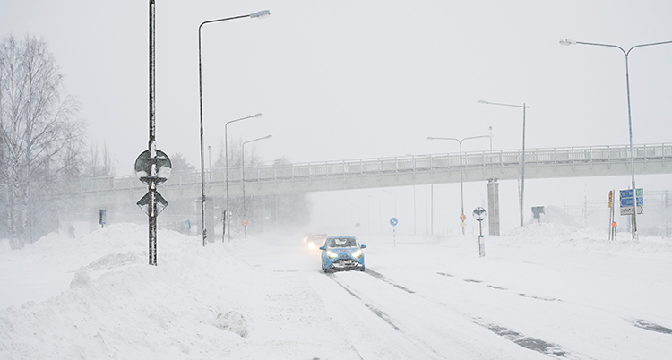 Vi ser en bilväg i staden Umeå. Några bilar kör på vägen. Det är snöigt på vägen och mycket snö bredvid vägen.