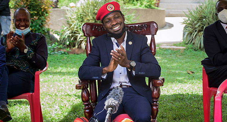 Bobi Wine sitter på en stol i sin trädgård. Han ler. Han har en röd basker på sig. På bordet framför honom ligger mikrofoner.