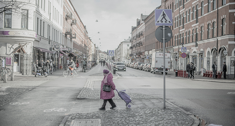 En kvinna med en väska på hjul går över ett övergångsställe i ett grått Malmö
