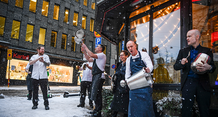 De står på en snöig trottoar utanför en restaurang, iklädda vita kockskjortor, och slår på kastruller
