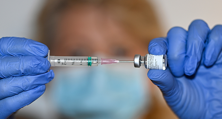 Två händer sprutar in vaccin från en liten flaska in i en spruta. Händerna har plasthandskar på sig.