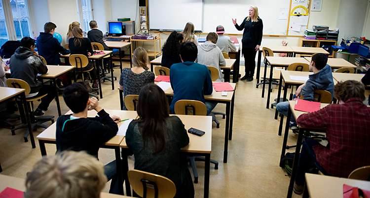 Ett klassrum. Bilden är tagen längst bak i klassrummet, fram mot tavlan. Elever sitter vid bänkar och lyssnar. Längst fram står en lärare och pratar.