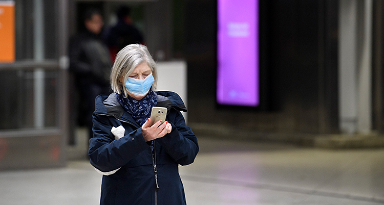 En kvinna står på en perrong. Hon väntar på en tunnelbana. Hon har ett munskydd på sig. Hon håller i en mobiltelefon.