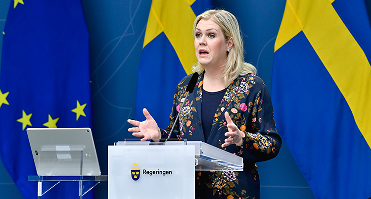 Lena Hallengren pratar för journalister. Hon står vid et litet bord och pratar i en mikrofon. Hon har långt ljust hår. I bakgrunden ser vi Sveriges flagga.