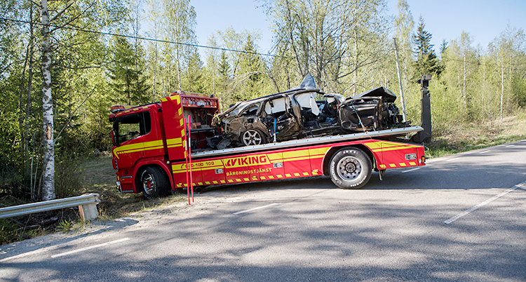 Bilden visar en landsväg. Det finns skog bredvid vägen. Himlen är blå. En röd bärgningsvbil håller på att backa upp på vägen. På flaket finns en personbil som är mycket skadad.