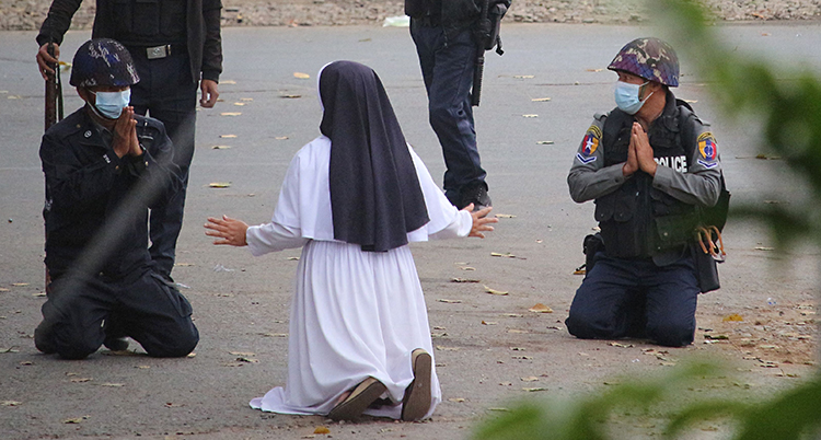 En nunna står på knä på marken. Vi ser henne bakifrån. Framför henne är fyra poliser. Två av poliser står också på knä.