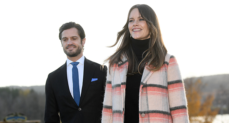 De är utomhus på landet. De går mot kameran. De är glada. Carl Philip är till vänster i kostym. Sofia är till höger i en kappa som är rosa, beige och grå.