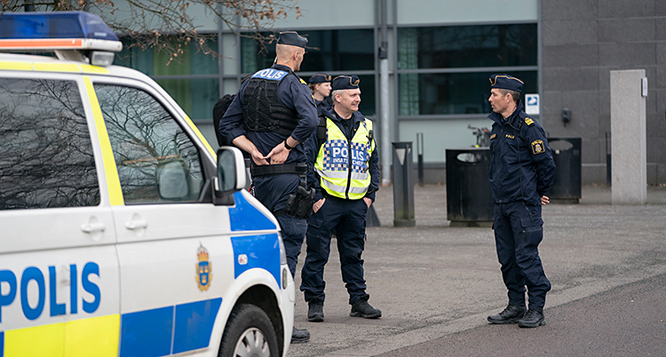 Tre poliser står och pratar med varandra. De står utanför en byggnad som är stor och grå. Bredvid poliserna är en polisbil.