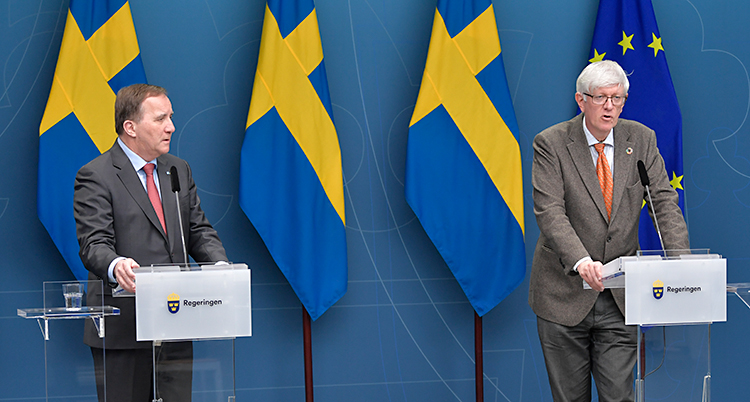 Löfven och Carlson står i ett blått rum. Bakom sig har de svenska flaggan och EUs flagga. De står vid varsitt litet bord och pratar i varsin mikrofon. De har kostym och slips.
