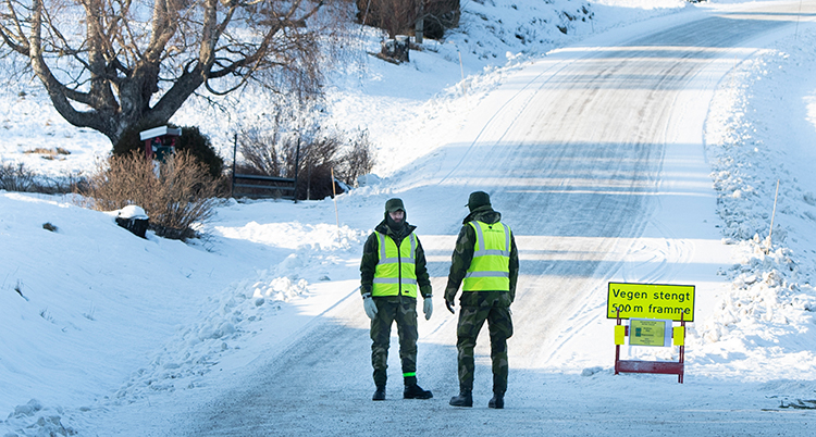 Två män i gula västar och gröna kläder står på en väg som är täckt av snö. De är troligen militärer. På en skylt står det på norska, Vegen stengt.