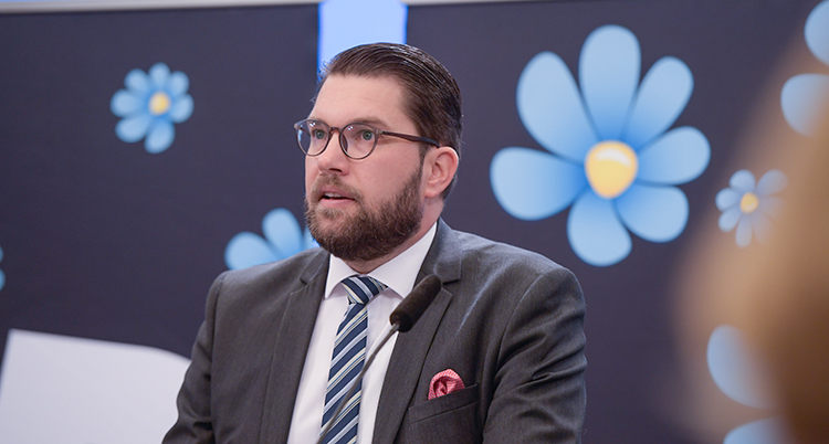 Åkesson ser allvarlig ut. Bilden visar överkroppen. Han pratar. Han har slips och kostym. Em röd näsduk sticker upp ur fickan. Bakom honom är partiets symbol - en blåklocka.