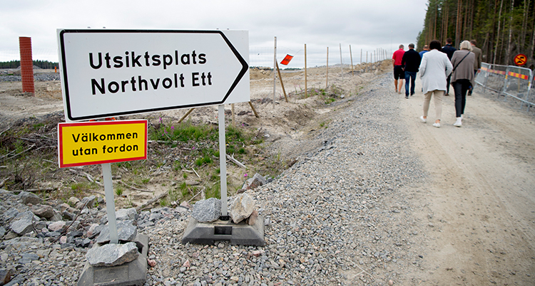 En vit skylt står intill en grusväg där människor går.