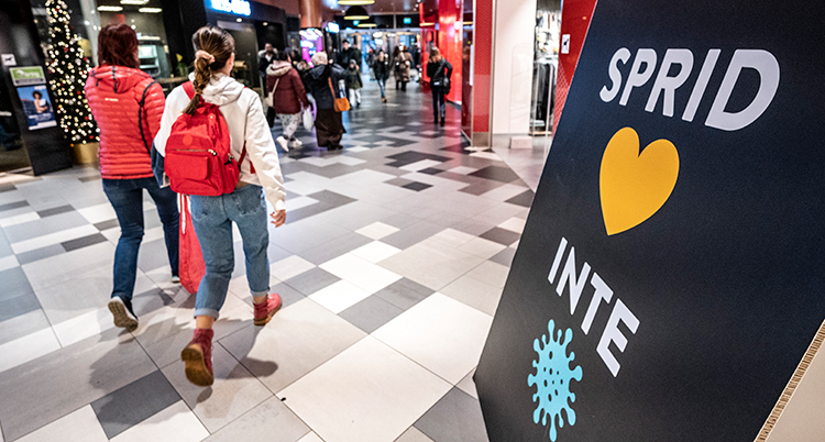 Två personer går bredvid varandra i ett köpcentrum. En skylt säger Sprid inte virus.