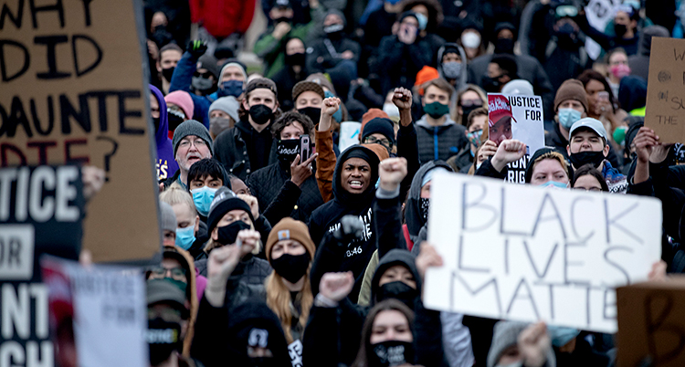 Många människor har samlats för att protestera. Flera sträcker upp en knuten näve. Många har plakat med sig. På ett av plakaten står det Black lives matter.