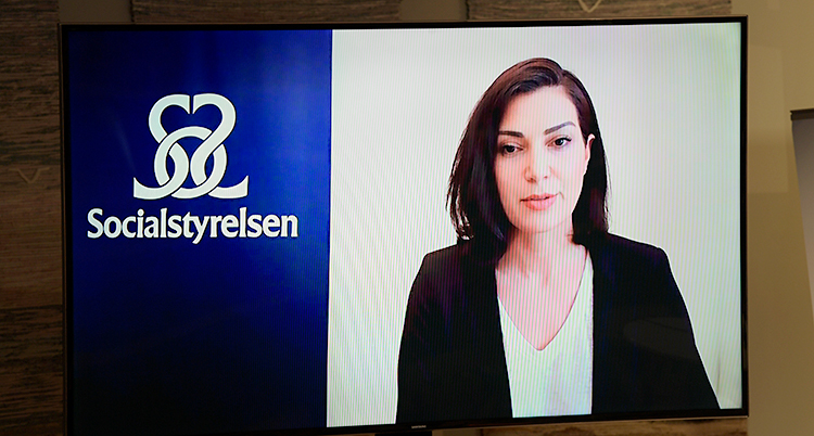 Vi ser en kvinna på en skärm. Hon pratar. Bredvid henne finns en skylt där det står Socialstyrelsen.