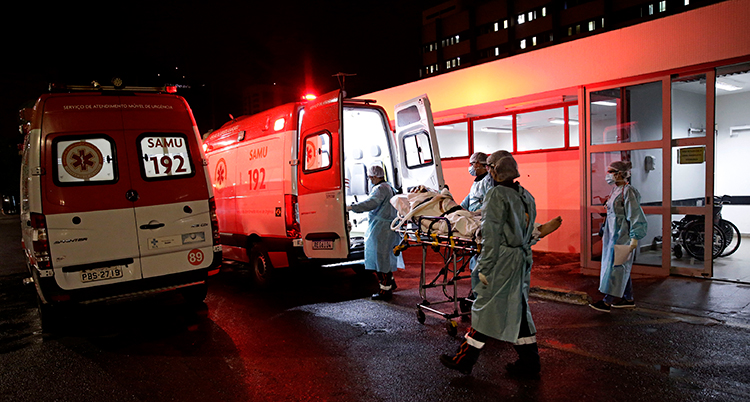 En röd ambulans har en öppna bakluckor. En människa bärs ut på en säng av flera personer. Det är mörkt ute.